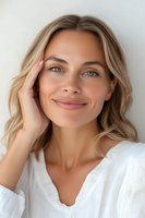 Young Caucasian woman with blonde wavy hair and natural makeup smiling gently while touching her face, wearing white blouse against light background. For beauty and skincare advertising.