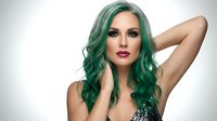 Young Caucasian woman with vibrant green wavy hair and bold makeup posing with hands in hair against neutral background for beauty and fashion concepts.