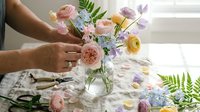 Florist arranging fresh spring bouquet with pink roses, ranunculus and fern in glass vase. Professional flower arrangement workshop for weddings and events.