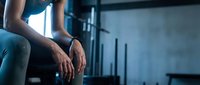 Young male athlete experiencing knee pain or injury while sitting in modern gym environment with equipment visible in background.