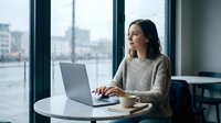 Young professional woman working on laptop in modern office space with large windows overlooking urban cityscape during daytime for business productivity.