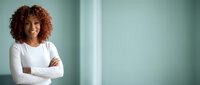 Confident African American businesswoman with curly hair in white shirt standing with crossed arms against mint green background for corporate branding.