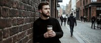 Young bearded Caucasian man in black sweater holding coffee cup while leaning against brick wall on busy urban street with blurred pedestrians walking.