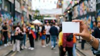 Hand holding blank white card in crowded urban street market with colorful storefronts and pedestrians, suitable for promotional messaging and advertising campaigns.