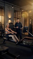 Personal trainer coaching mature blonde woman on rowing machine in modern gym with warm lighting and glass partitions for fitness motivation.