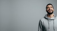 Middle-aged man with dark beard and hair, Mediterranean ethnicity, wearing gray hoodie, smiling and looking upward against neutral gray background. Studio portrait for lifestyle, casual wear, and apparel marketing.