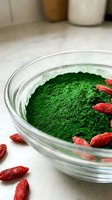 Vibrant green matcha powder in glass bowl with red goji berries scattered around, wellness and superfood ingredients for healthy nutrition and beverage preparation.