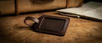 Brown leather luggage tag with clear window on wooden surface next to vintage suitcase for travel identification and baggage organization accessories.
