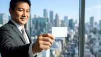 Smiling Asian male professional in gray business suit holding blank white business card with urban skyline visible through office window behind him.