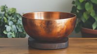 Handcrafted copper bowl with rich patina finish on wooden stand, surrounded by green plants on natural wood surface for kitchen decor and serving.