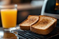 Fresh toasted bread slices on electric toaster grill with glass of orange juice in background, morning breakfast setting on wooden table surface.