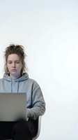 Young Caucasian woman with messy hair working on laptop wearing gray hoodie against white background. Tired student or remote worker concept.