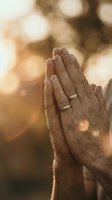 Hands pressed together in prayer position with wedding rings against warm golden bokeh light background for spiritual meditation and mindfulness concepts.