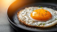 Sunny side up fried egg with crispy edges and black pepper seasoning in cast iron pan, bright orange yolk and white protein on dark background.