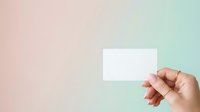 Person with medium skin tone holding blank white business card against soft pastel gradient background in pink and mint tones, suitable for branding and corporate identity mockups.
