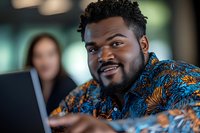 Young African American businessman smiling while working on laptop in modern office setting, wearing colorful patterned shirt. Professional workplace environment.