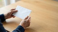 Senior adult with medium skin tone holding blank white envelope at wooden table, suitable for mail, correspondence, business communication, and administrative concepts.