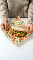 Hands holding a fresh veggie sandwich wrapped in floral beeswax reusable wrap on white table, eco-friendly zero waste food storage concept.