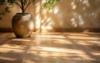 Sunlight and tree shadows cast dramatic patterns on wooden floor near ceramic vase, creating zen atmosphere with natural dappled light effect. Minimalist home decor.