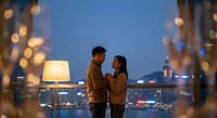 Young Asian couple embracing on city rooftop terrace at twilight with bokeh lights and urban skyline background for romantic evening date concept.