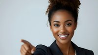 Young African American businesswoman with curly hair in bun wearing black blazer pointing and smiling confidently against light background, professional corporate portrait.