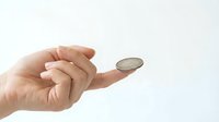 Hand holding single coin on fingertip against clean white background, symbolizing financial decisions, savings, investment choices, and monetary value concepts.