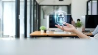 Hand holding glowing digital security lock icon in modern office workspace representing cybersecurity data protection and network safety technology.