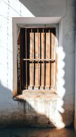 Rustic wooden window with vertical bars in white stucco wall, dramatic natural sunlight casting shadows creates moody architectural detail.