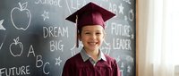 Young Asian girl in burgundy graduation cap and gown smiling in classroom with chalkboard featuring educational symbols and motivational words.