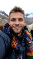 Smiling Caucasian man in winter jacket and colorful knitted scarf taking selfie outdoors with snowy mountains in background, authentic travel lifestyle portrait.