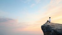 Young Asian man sitting alone on cliff edge overlooking peaceful sunset sky with soft pastel colors, representing solitude, meditation and life achievement.