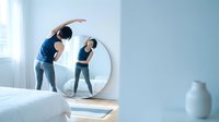 Young Asian woman doing side stretch exercise in front of circular mirror in bright modern bedroom for morning fitness routine and healthy lifestyle.
