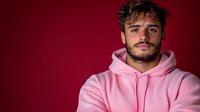 Young bearded man wearing pink hoodie against dark red background, looking directly at camera with confident expression. Modern casual fashion portrait.