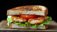 Fresh grilled chicken sandwich with crisp lettuce and juicy tomato slices on toasted bread, served on rustic wooden board against dark background.