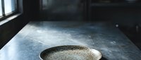 Young adult male with light skin tone, wearing casual attire, pouring sand or granular material into a metal bowl on a dark industrial surface, suitable for construction, material handling, or craft project concepts.