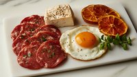 Fresh sliced beef carpaccio with sunny side up egg, dried orange slices, butter and microgreens served on white marble plate, gourmet appetizer presentation.