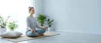 Young woman practicing meditation in lotus position on yoga mat in bright minimalist room with plants. Peaceful mindfulness and wellness lifestyle concept.