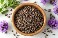 Dried black tea leaves in wooden bowl with fresh purple chive blossoms and green herbs on white background, overhead view for food and beverage photography.
