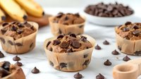 Chocolate chip muffins with dark cocoa drops on marble counter, fresh bananas and wooden spoon, homemade pastry closeup view in natural light.