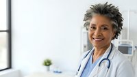 Smiling African American female doctor with stethoscope in white coat standing in modern medical office with natural light from window.