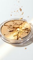 Broken golden face powder compact with scattered fragments and particles floating in air on white background for beauty and cosmetics concepts.