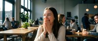 Young professional woman laughing with hands covering mouth in modern office workspace with colleagues working in background during break time.