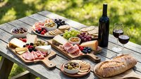 Outdoor charcuterie board spread with cured meats, cheeses, crackers, fresh fruits, olives, and wine on wooden table in garden setting for entertaining and gatherings.