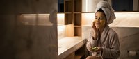 Young Asian woman in bathrobe and towel relaxing in modern bathroom after shower, holding skincare product for morning beauty routine and self-care ritual.