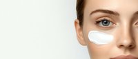 Close up of woman's face with white moisturizing cream or under eye patch on cheek against light background, demonstrating skincare and beauty treatment application.
