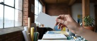 Hand holding blank white business card in modern loft office with brick walls and large windows. Mockup template for branding and corporate identity design.