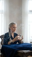Mature Caucasian woman fortune teller with braided hair holding crystal ball at table with mystical smoke in elegant room for spiritual guidance sessions.