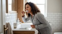 Young African American woman brushing teeth in modern bathroom with white subway tiles. Morning dental hygiene routine in contemporary home interior.