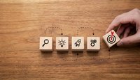 Hand placing wooden blocks with business icons showing search, idea, rocket, gears and target symbols on wood surface for strategy planning.