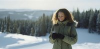 Young woman with curly hair smiling in winter landscape, wearing green parka and gloves against snowy mountain backdrop with evergreen forest.
